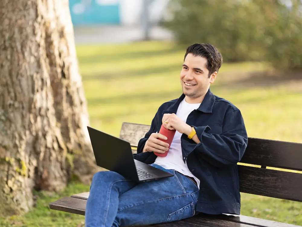 Un dipendente Henkel è seduto su una panchina in un parco e lavora al suo laptop. 