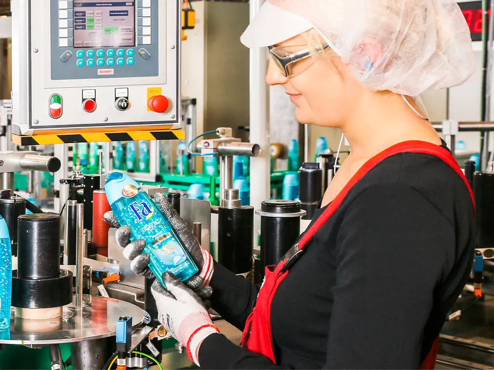 A female employee stands next to the assembly line inspecting a Fa bottle.
