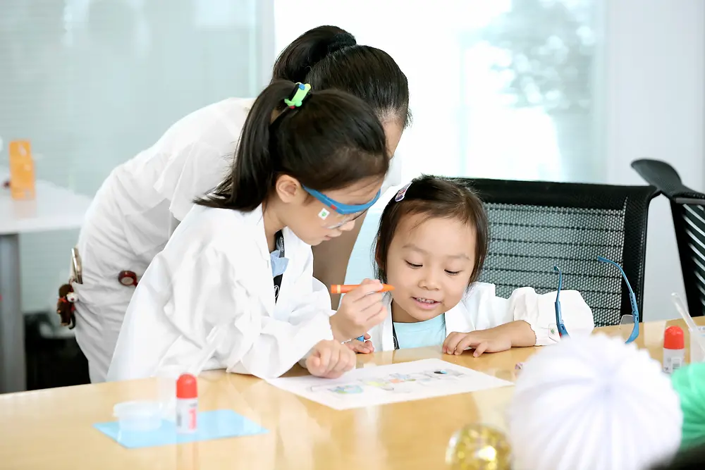 Due bambine e una donna con camici da laboratorio colorano un disegno su un tavolo