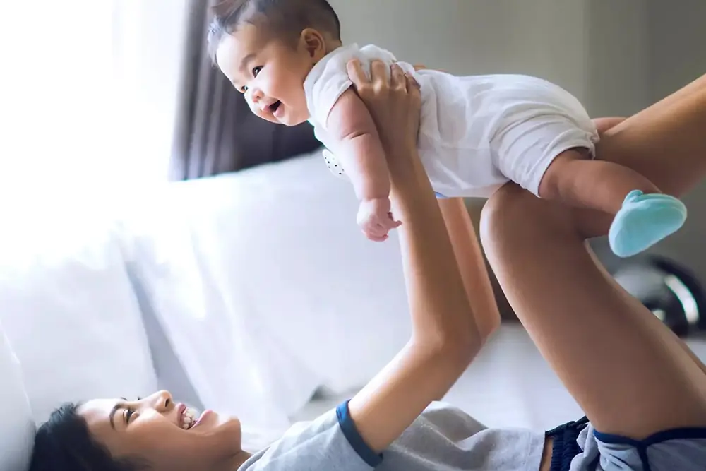 Una madre solleva e gioca con un bambino sorridente in un ambiente domestico luminoso.