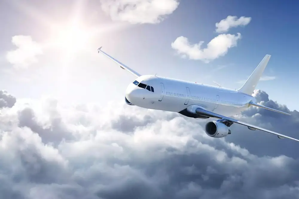 Un aereo passeggeri bianco vola sopra soffici nuvole in un cielo luminoso, con la luce del sole dall’alto a sinistra.