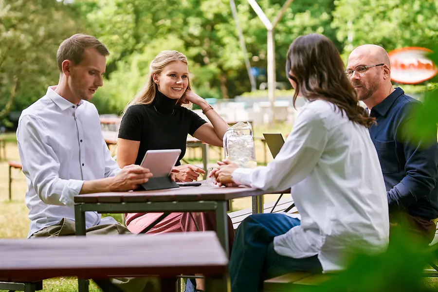 Gruppo di colleghi seduti insieme all'aperto, che discutono di lavoro con tablet e laptop, a rappresentare la collaborazione e una cultura aperta e orientata al team in Henkel.