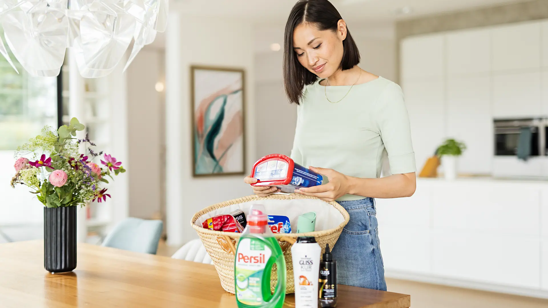 Donna con un cesto sul tavolo pieno di prodotti Henkel come Persil, Gliss, Bref, Somat, taft e altri.