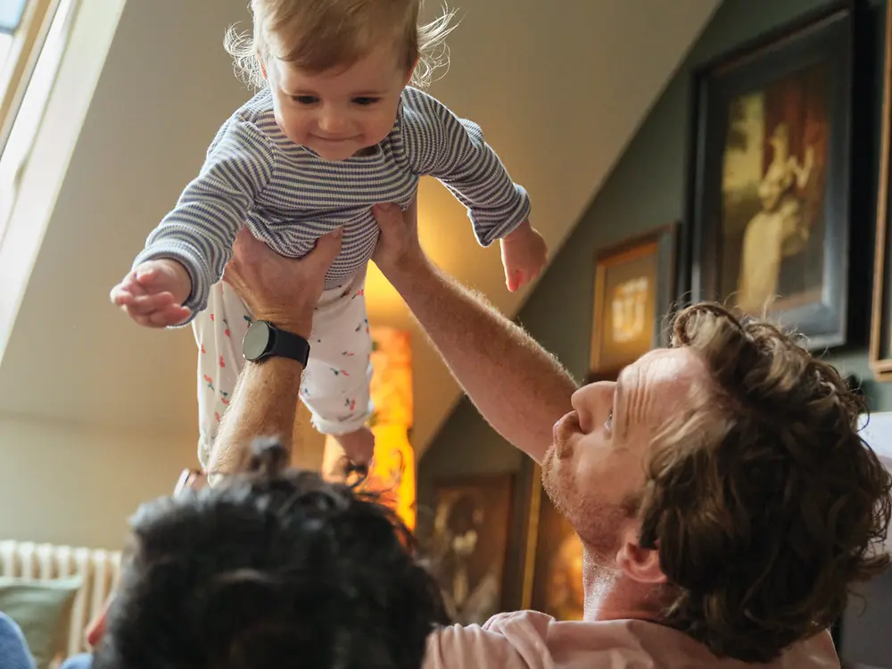Due papà sorridono mentre tengono in braccio il loro bambino a casa, a simboleggiare inclusività e la politica di congedo parentale attenta alle famiglie di Henkel.