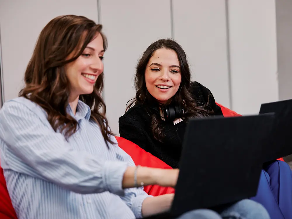 Due persone sedute su colorati pouf in un moderno ambiente d'ufficio, entrambe al lavoro con un computer portatile. Sembrano discutere dei benefit per i dipendenti Henkel mentre collaborano a un progetto.