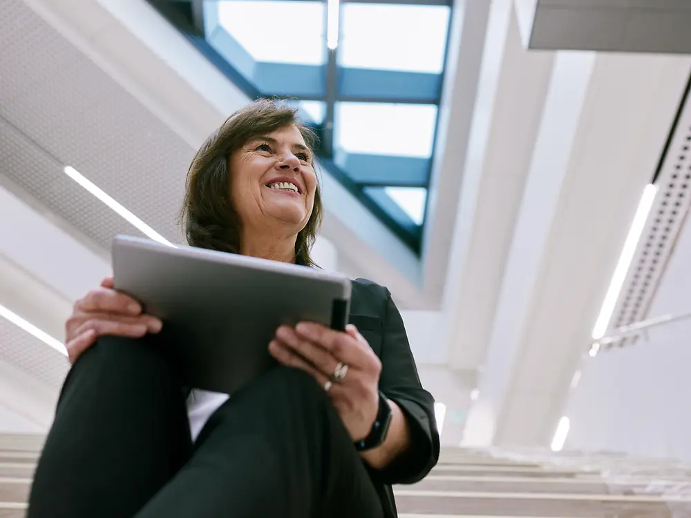 Una persona seduta sulle scale interne di un edificio moderno, con un tablet in mano. Lo sfondo mostra un design architettonico luminoso e aperto, caratterizzato da ampie finestre e lucernari, che crea un’atmosfera pulita e professionale.