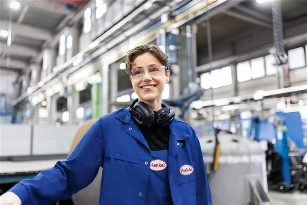Un dipendente Henkel sorridente con camice da lavoro blu e cuffie di sicurezza, in piedi in un capannone di produzione industriale, che rappresenta competenza e innovazione nella produzione.