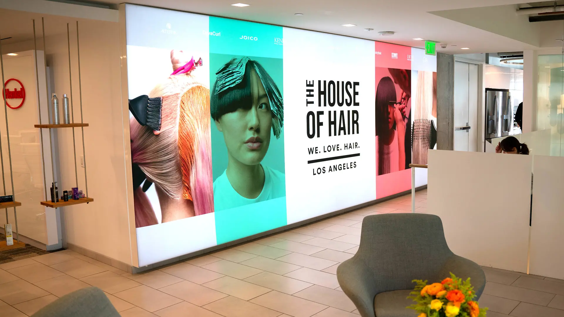 Una moderna hall con un luminoso display a parete con grandi pannelli tematici a colori e la scritta "THE HOUSE OF HAIR – WE. LOVE. HAIR. – LOS ANGELES.” Lo spazio comprende poltrone grigio chiaro, un tavolino con una vivace composizione floreale e un design pulito e minimalista con pavimento piastrellato e divisori bianchi.