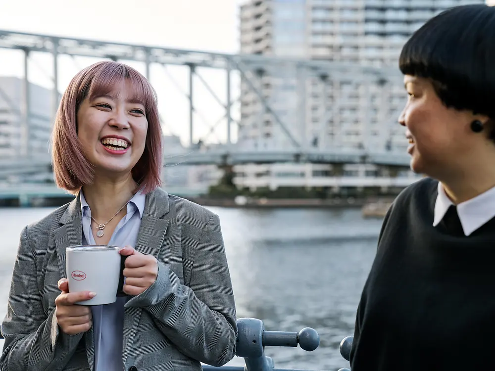 Due persone conversano informalmente vicino a una balaustra sul lungofiume; una di loro tiene una tazza da caffè con il marchio Henkel. Sullo sfondo si vedono un ponte e alcuni edifici cittadini, a rappresentare la cultura del lavoro flessibile di Henkel.