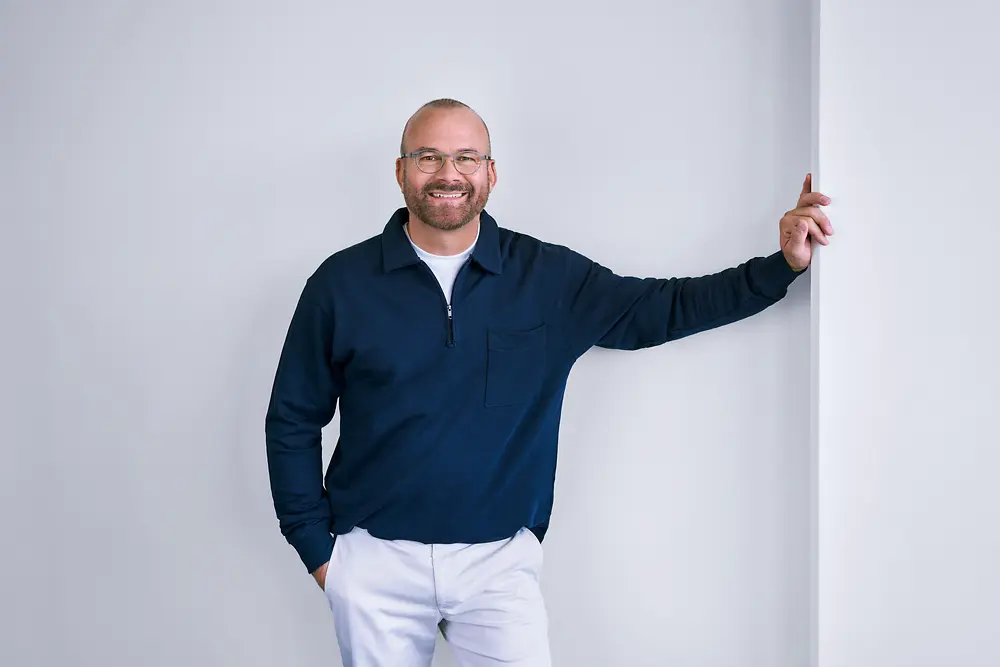 Professionista esperto di Henkel, in piedi con sicurezza davanti a una parete bianca in un ambiente moderno, con una camicia scura a maniche lunghe e pantaloni di colore chiaro.