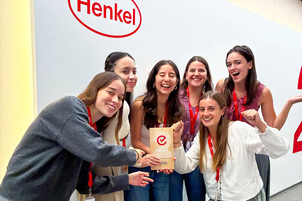 Un gruppo di sei persone è in piedi davanti a una parete con il logo Henkel. Indossano cordini rossi e abiti casual e sono riunite attorno a una targa di riconoscimento. L'ambiente è uno spazio aziendale interno con pareti chiare e pavimento grigio. Il gruppo posa con entusiasmo, mettendo in risalto il premio al centro.