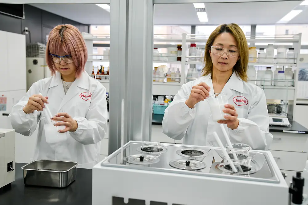 Due persone con camici bianchi Henkel lavorano in un moderno laboratorio. Manipolano strumenti scientifici e contenitori su un banco da lavoro, mentre sullo sfondo sono visibili attrezzature e scaffali con materiali di laboratorio.