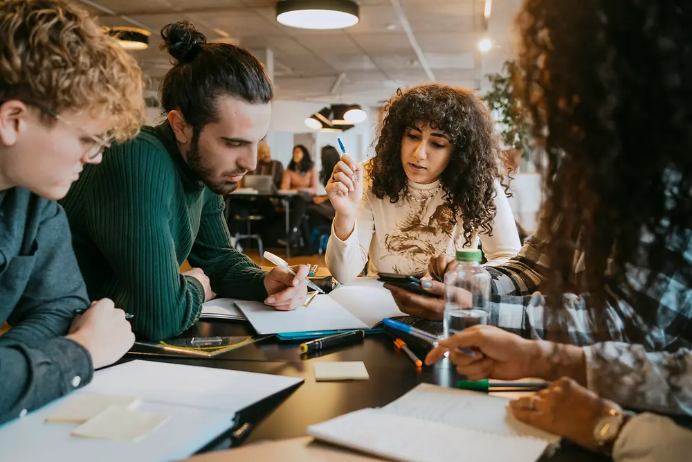 Un gruppo di persone riunite attorno a un tavolo in un moderno ambiente interno di lavoro collabora su documenti, quaderni e dispositivi digitali. Sul tavolo sono presenti penne, fogli e post-it, mentre il team è impegnato nel confronto, nella condivisione di idee e nella presa di appunti.