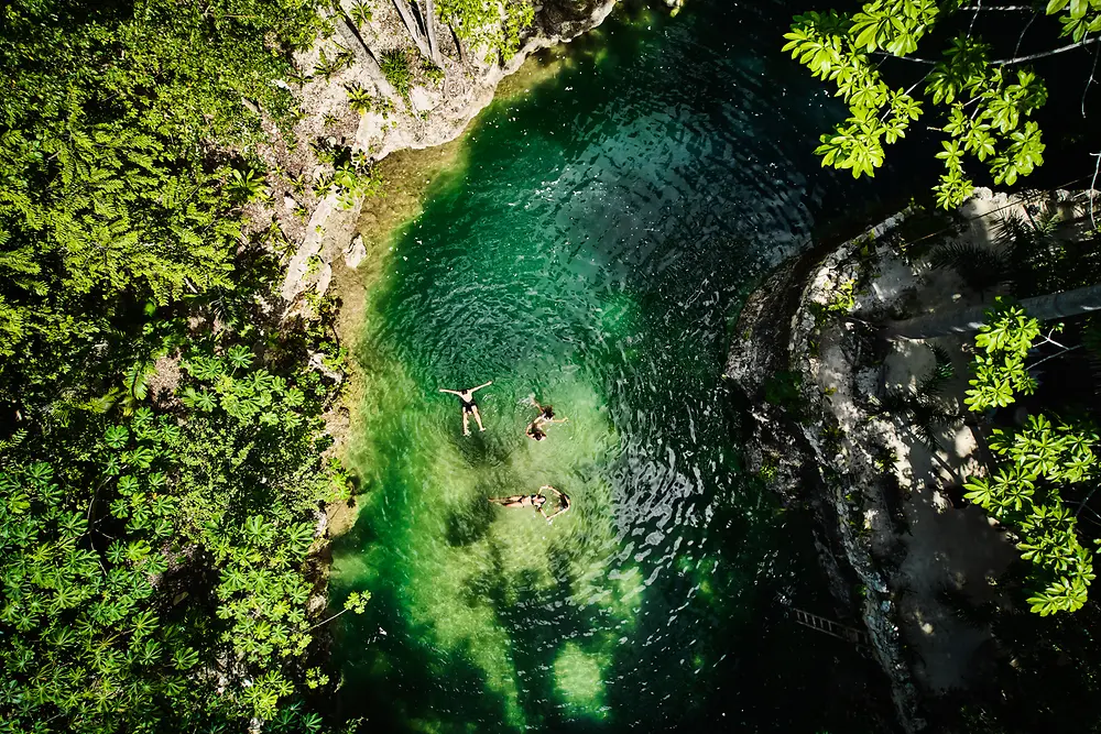 Vista aerea di una piccola piscina naturale circondata da una fitta vegetazione verde, con tre persone che galleggiano nell’acqua limpida color smeraldo.