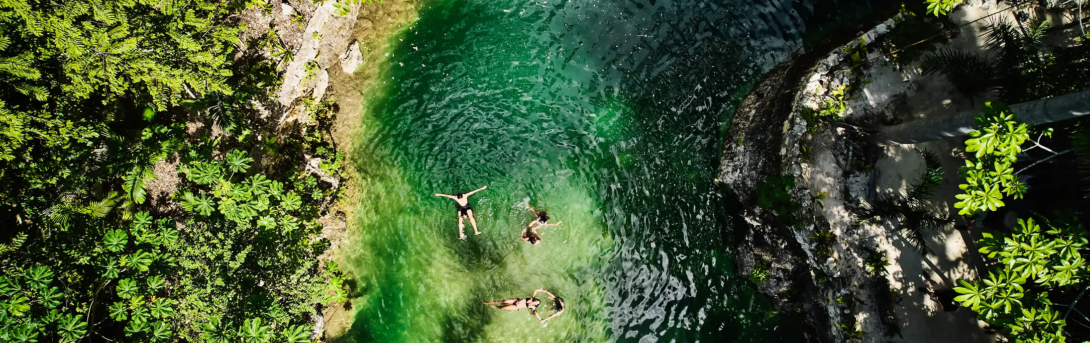 Vista aerea di una piccola piscina naturale circondata da una fitta vegetazione verde, con tre persone che galleggiano nell’acqua limpida color smeraldo.