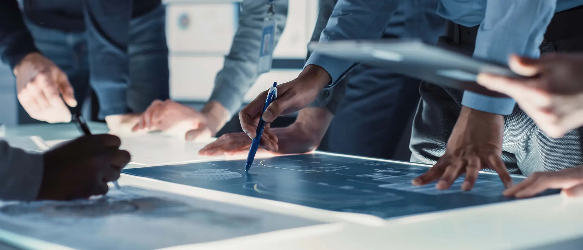 Vista ravvicinata di più persone riunite attorno a un ampio tavolo luminoso, impegnate nell’analisi e nell’annotazione di progetti tecnici mediante l’utilizzo di penne e dispositivi digitali. Un contesto che richiama collaborazione, precisione e innovazione.