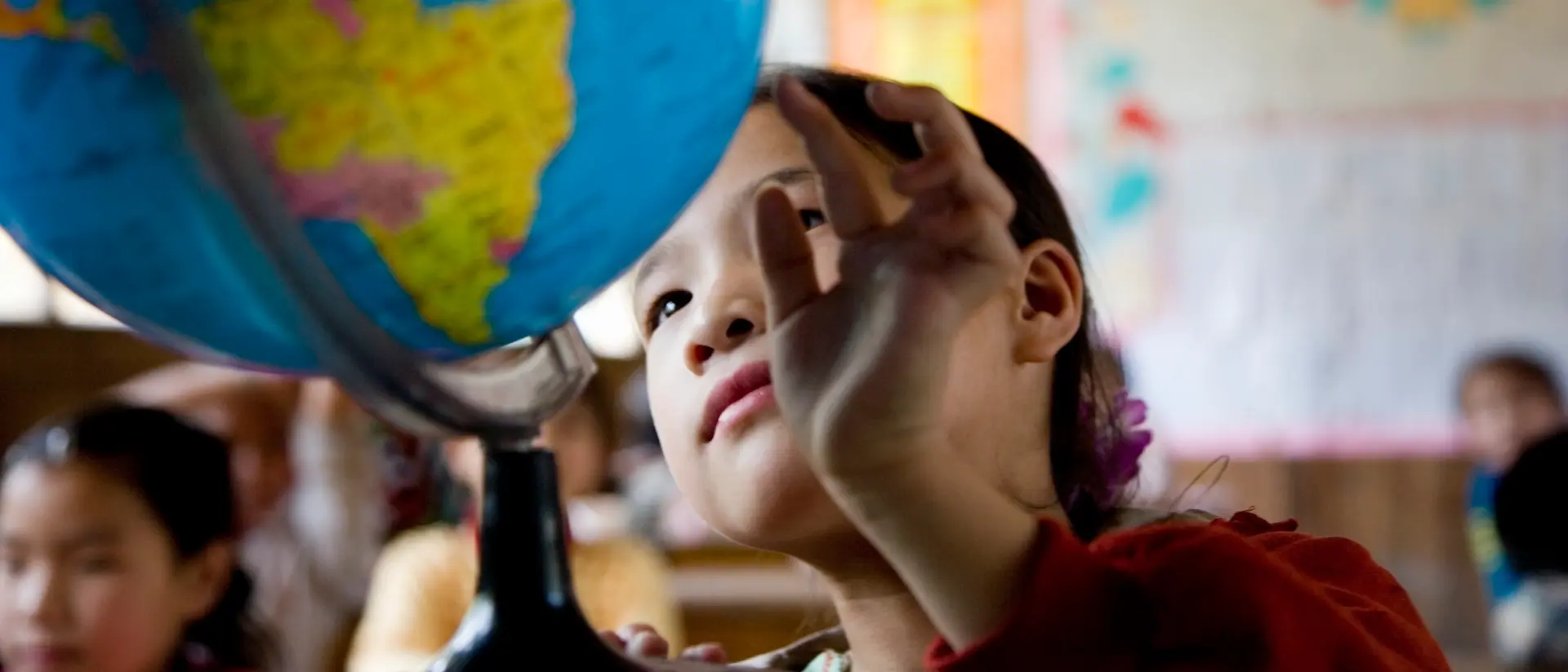 Un bambino, in un’aula scolastica, si protende verso un grande mappamondo appoggiato su un banco; sulle pareti sono visibili mappe colorate e materiali didattici. Un contesto che richiama l’apprendimento, la curiosità e la scoperta del mondo.