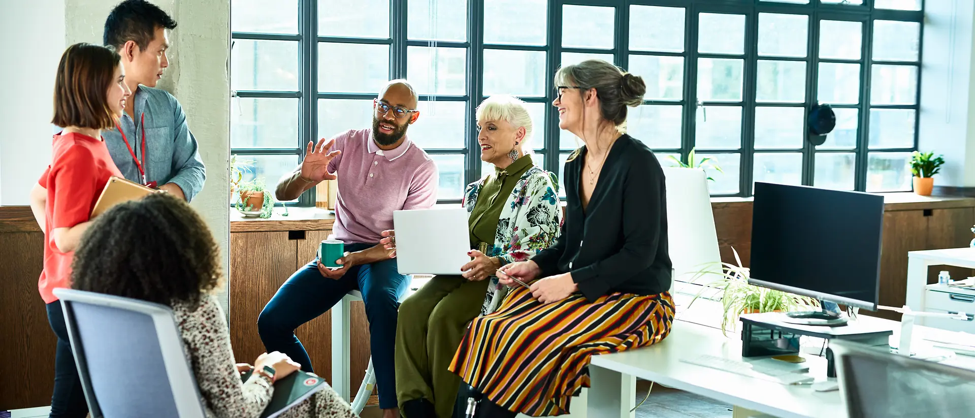 Un piccolo gruppo di persone, sedute e in piedi, dialoga in un ufficio luminoso e open space, in prossimità di postazioni di lavoro e ampie finestre. Un contesto che favorisce il confronto, la collaborazione e la condivisione di idee.