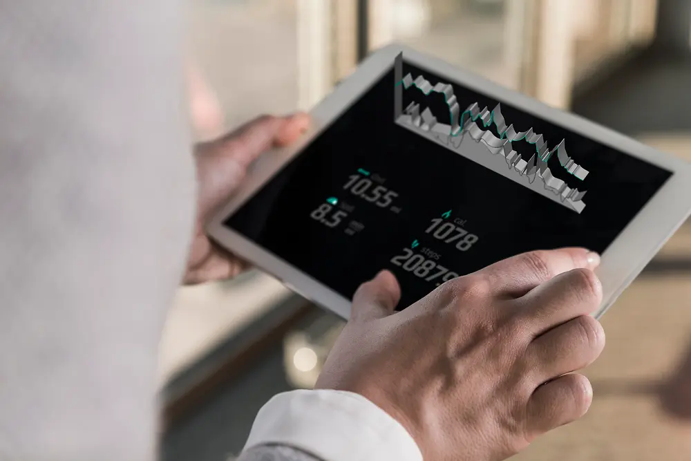 Una persona osserva un tablet che visualizza grafici digitali e dati numerici, ripresa da dietro la spalla in un ambiente indoor luminoso.