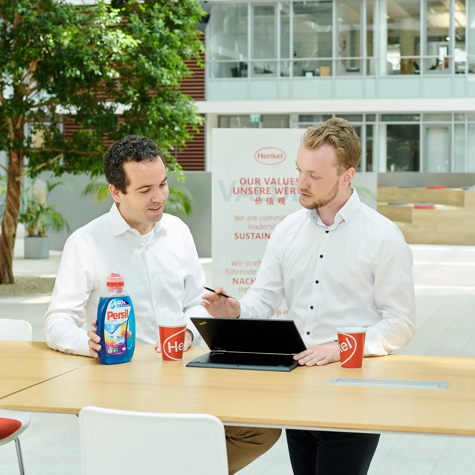 Due uomini in piedi dietro un tavolo con un flacone di Persil e un laptop